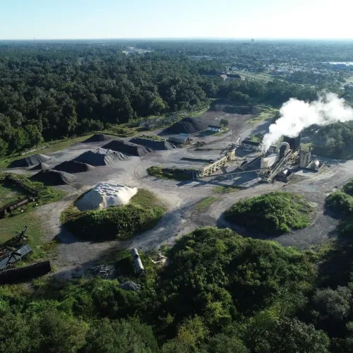 Drone image of an active asphalt facility with visible stockpiles, smoke-emitting machinery, and surrounding forested areas supporting industrial operations