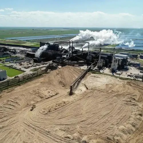 Aerial view of an sugar plant adjacent to a wooded area and pond, with visible stockpiles and facility infrastructure