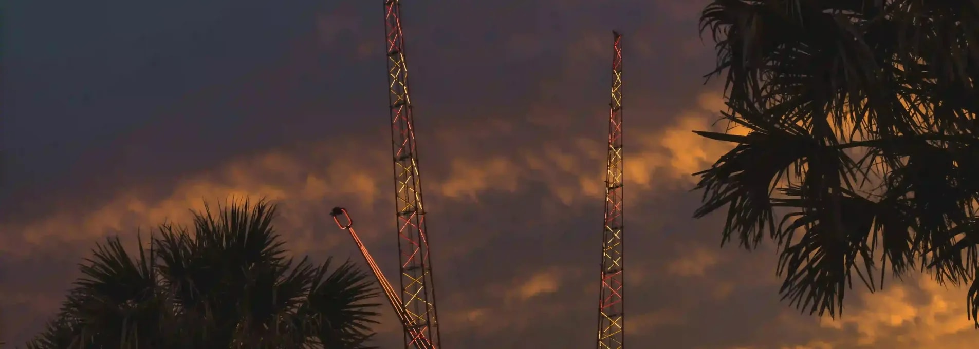 Amusement ride towers lit in red and yellow lights silhouetted against a dramatic sunset sky with palm trees