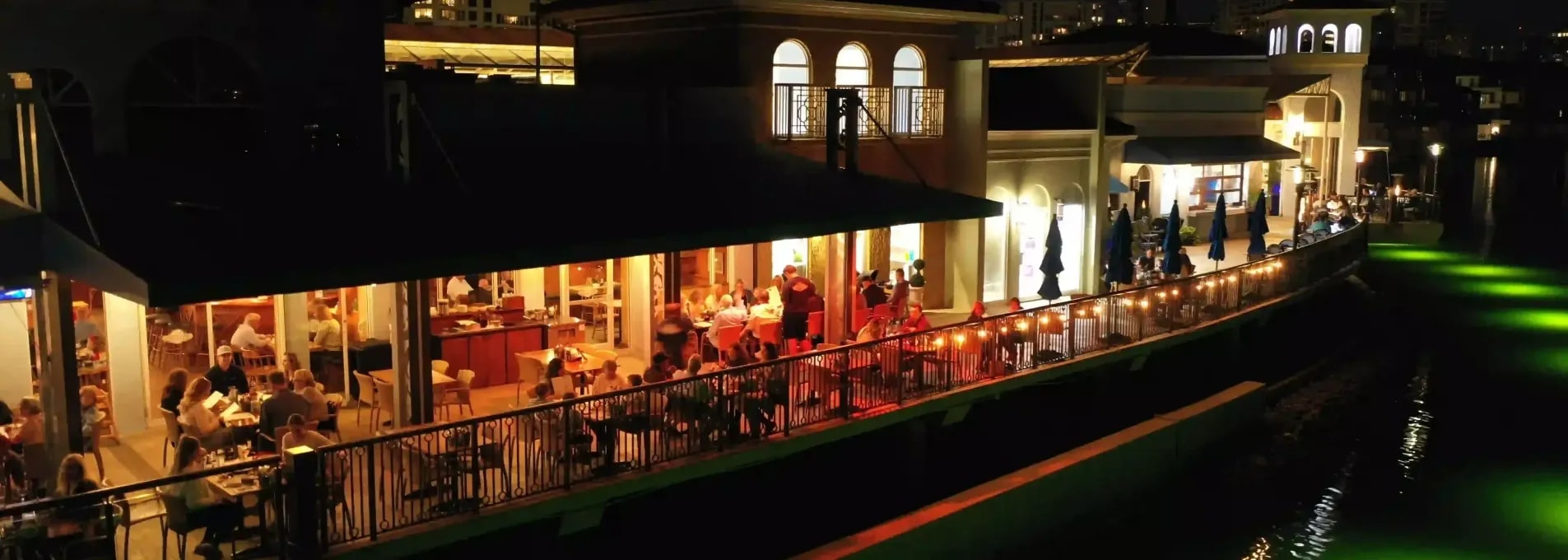 Evening drone view of open-air dining terrace beside a canal, with lights reflecting on the water and buildings in the background