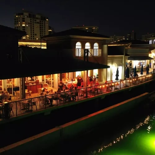 Evening drone view of open-air dining terrace beside a canal, with lights reflecting on the water and buildings in the background