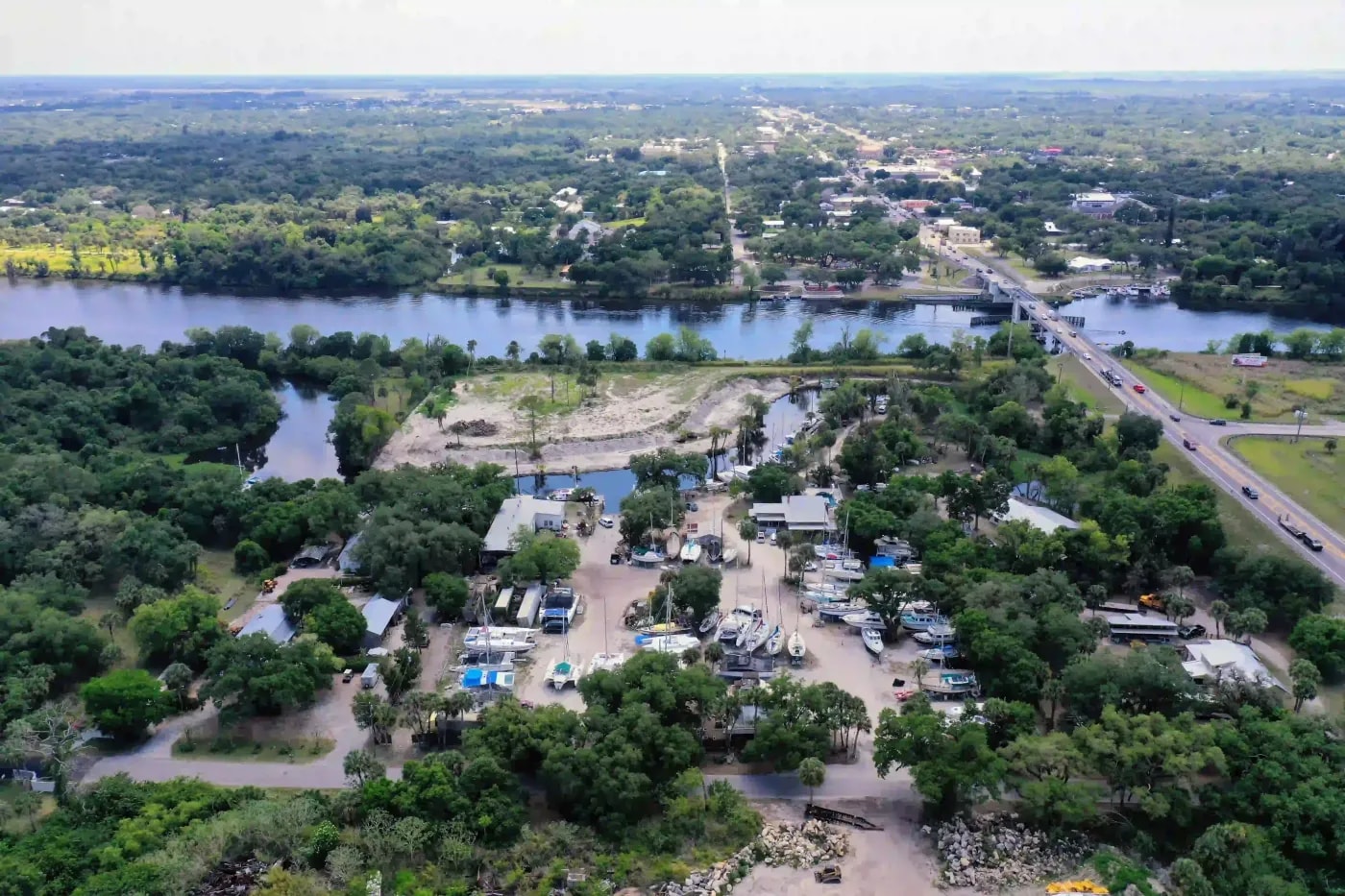 Aerial image of boatyard and marina near a wooded area with access to a wide river and a distant bridge crossing