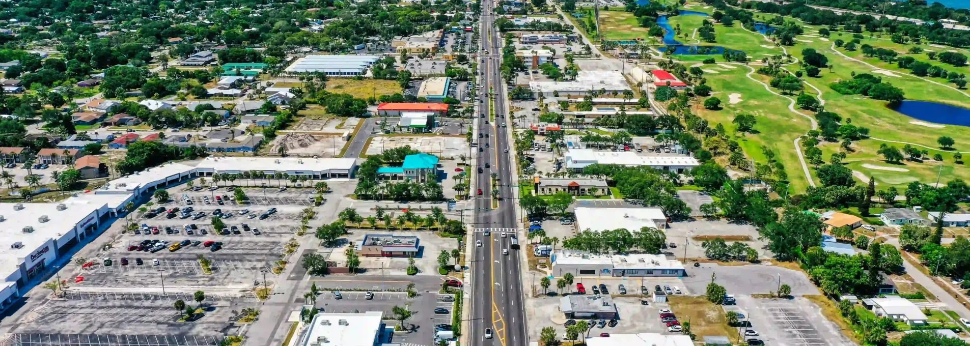 Aerial photo of a commercial boulevard flanked by retail centers, a golf course, and a coastal bridge in the distance